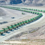 20 lines and 100 number of mining dump trucks located middle of the picture and left and right side is a mountains. the huge truck in middle of them has green lights and cargo and body that resembles like the robots in the Transformers movie
