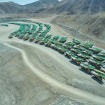 20 lines and 100 number of mining dump trucks located middle of the picture and left and right side is a mountains. the huge truck in middle of them has green lights and cargo and body that resembles like the robots in the Transformers movie