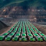 100 number of mining dump trucks located middle of the picture and left and right side is a mountains. the huge truck in middle of them has green lights and cargo and body that resembles like the robots in the Transformers movie