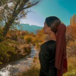 یک خانم معلم زیبا پشت به تصویر با روسری ولباس بلند در فضایی پر از درخت و رودخانه و کوه ودرختان رنگ پاییزی