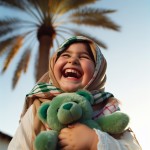 دختر 9 ساله تپل با چادر عربی که در دست آن یک عروسک خرسی سبز قرار دارد و دختر در حال خندیدن است. پشت آن یک نخل قرار دارد که نور خورشید به آن تابیده است.