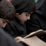 پسر12 ساله ایرانی در حال مطالعه زیارت عاشورا با لباس مشکی در کنار دیگر کودکان ایرانی و عراقی در یکی از موکب های مسیر پیاده روی اربعین