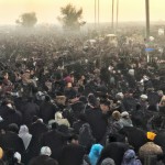 عکس سردار سلیمانی را که در حال پیاده روی اربعین است و در میان انبوه جمعیت در مسیر نجف تا کربلا هست و همه زایران در این مسیر در حال حرکت هستند و عمود ۸۰۰ در جاده را نشان میدهد. موکب های اطراف جاده در حال پخش چای و شربت و غذا هستند.