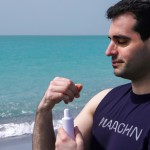 یک مرد جوان خوشتیپ با سبیل‌های کلفت کنار ساحل زیبای خلیج فارس در حال استفاده از کرم ضدآفتاب و تبلیغ و معرفی محصولات آرایشی بهداشتی مراقبتی برند MND اسم برند محصولات کاملا واضح و در معرض دید باشد