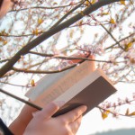 یک تصویر با جزئیات از دختری که در کنار درختان بهاری کتاب میخواند