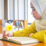 عکس دختر دانشجو چادری در کتابخانه نشسته روی میز در حال مطالعه و نیم رخ به همراه گل نرگس روی میز