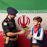 دانش آموز پسر ایرانی در حال دادن گل قرمز به پلیس مرد ایرانی در کنار پرچم ایران