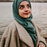 تصویر یک دختر 22 ساله ایرانی که شاداب هم هست و حجاب کامل دارد.صورت جذابی دارد و رنگ چشم هایش قهوه ای است.  رنگ لباسش فیروزه ای است و روی مانتوی او ، طرح های اسلیمی وجود دارد. او در کنار زاینده رود ایستاده است. موهای او معلوم نیست .