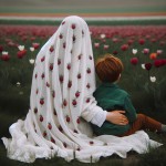یک مادر محجبه نشسته. پشتش به دوربین. چادر سفید روی سرش با طرح دونه‌های انارِ پراکنده. پسر ۴ ساله‌ش پشتش به دوربین. از بغل به مامان تکیه داده. موهای پسره خرمایی‌‌رنگ با ژاکت سبز و شلوار قهوه‌ای. دست مادر روی سر پسره. زیرشون چمن و روبه روشون منظره‌ای از یک دشت لاله