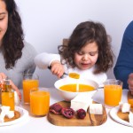 i want an image of a breakfest table that has feta cheese and eggs and jam on it and i want a family of three(wife,husband,kids) sitting at the desk and happily eating breakfest and the kid is holding a transparent glass of orange juice
