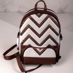 The image shows a white backpack with a brown and white zigzag pattern on its strap. The backpack features a quilted design with a front pocket and a metallic ring near the top.