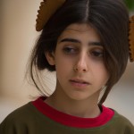 تصویر یک دختر بچه هفت ساله ایرانی که مانتو وشلوار پوشیده چشمان مشکی دارد وموهای او کمی از مقنعه او بیرون است