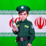 عکس واقعی یک پسر ۶ ساله با لباس سبز پلیس ایران با بیسیم به دست و پس زمینه مات پرچم ایران بساز