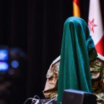 تصویر یک مرد ایرانی با لباس جنگی در حالی صورت خود را با چفیه پوشانده با پس  زمینه  یک کنفرانس خبری
