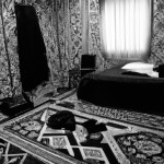 یه عکس بدون رنگ از یه اتاق که روبه رو یه پنجره هست و سمت راست یه تخت و سمت چپ کمد هست و روی زمین فرش تبریزی پهن هست و روی فرش یه چمدون ایرانی هست و روی تخت و فرش لباس و کتاب ریخته