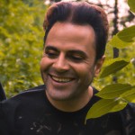عکس از خاننده معروف ایرانی حامیم درحال لبخند زدن و چشم هایی که از عشق پر شده و عاشق است در یک جنگل و در حالی که یکم سرش رو کج کرده