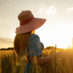 دختری کنار مزرعه گندم با کلاه لبه دار در حال نگاه کردن به غروب افتاب